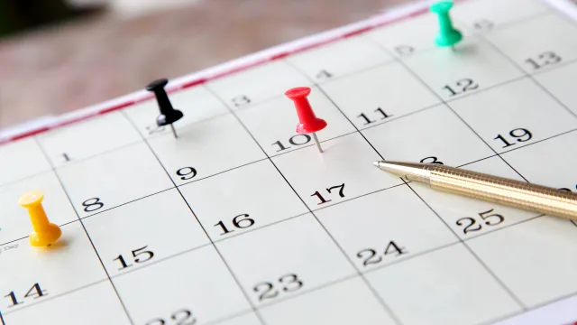 An image of a calendar with a pen and colourful pushpins resting on some of the dates. 