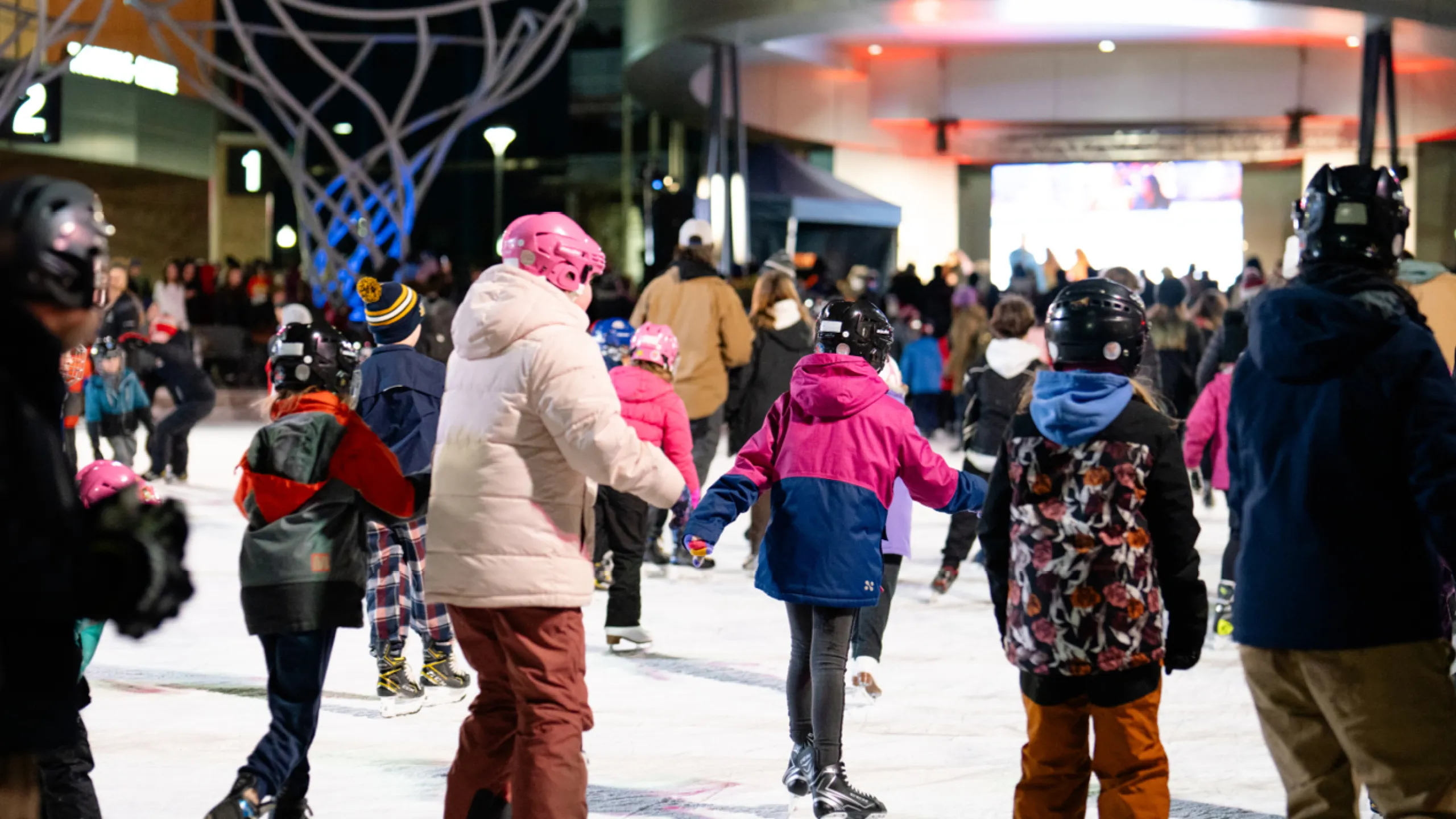 Crowd of people ice skating at night on a downtown outdoor rink.