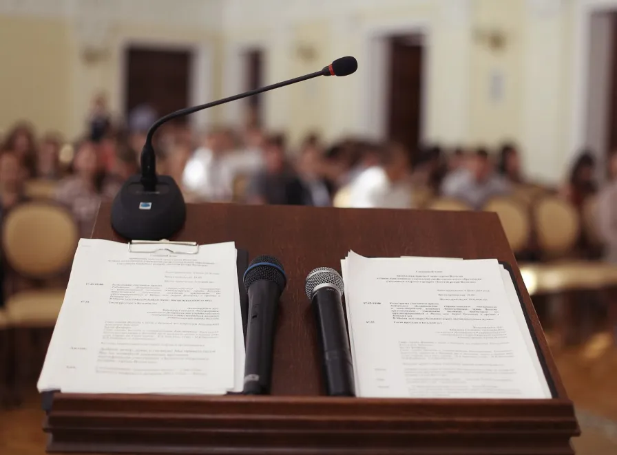 Podium with papers and two handheld microphones; gooseneck mic facing a blurred audience.