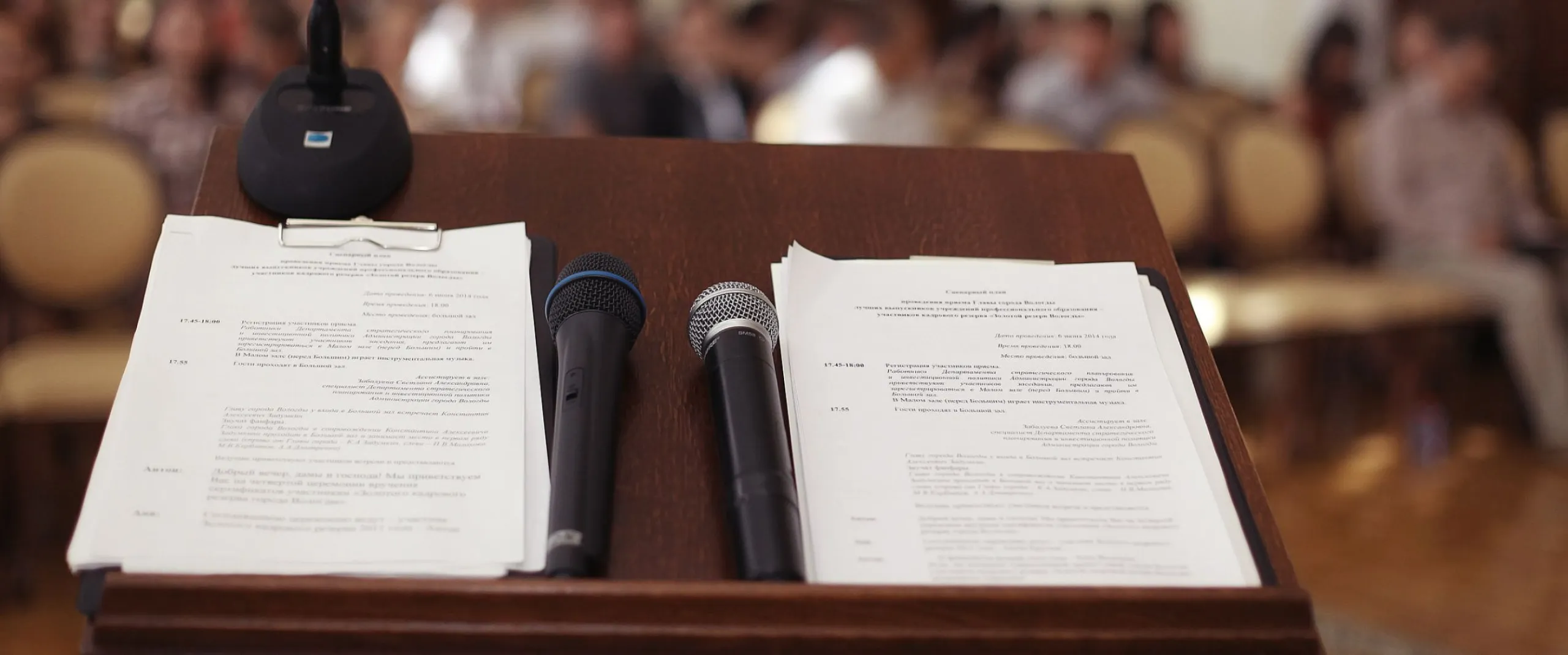 Podium with papers and two handheld microphones; gooseneck mic facing a blurred audience.